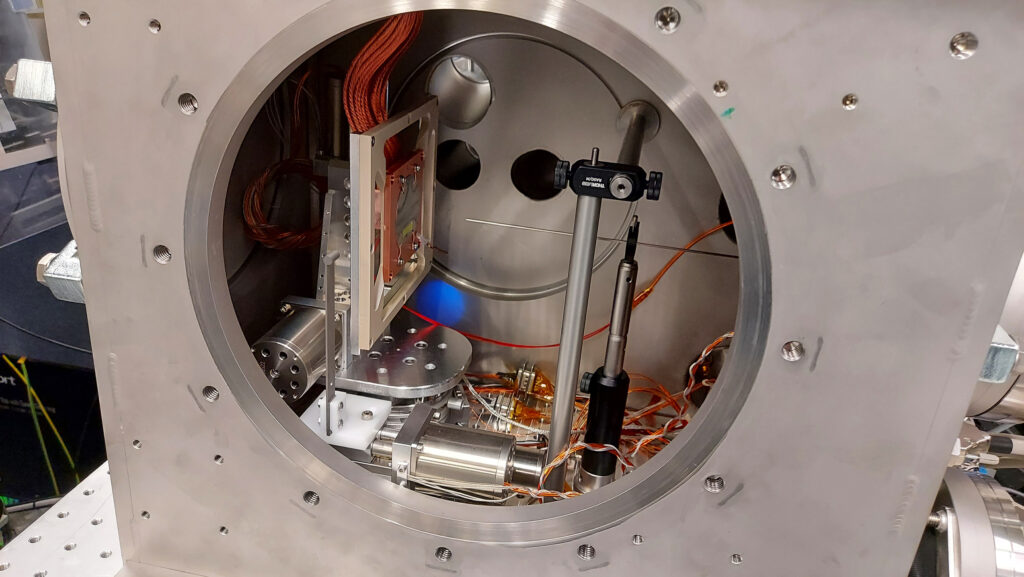 Custom-built vacuum chamber setup for the experiment: The round wafer, which is vertically mounted on a copper surface on the left, is coated with a platinum layer and was cooled down to -173 °C (100 K). Water vapor was released from the thin steel nozzle in the center, directly facing the wafer, which resulted in the formation of amorphous ice on the wafer. This ice was then excited by an optical laser and probed by X-rays coming in through the half-shadowed dark hole in the back of the chamber. (photo/©: European XFEL)